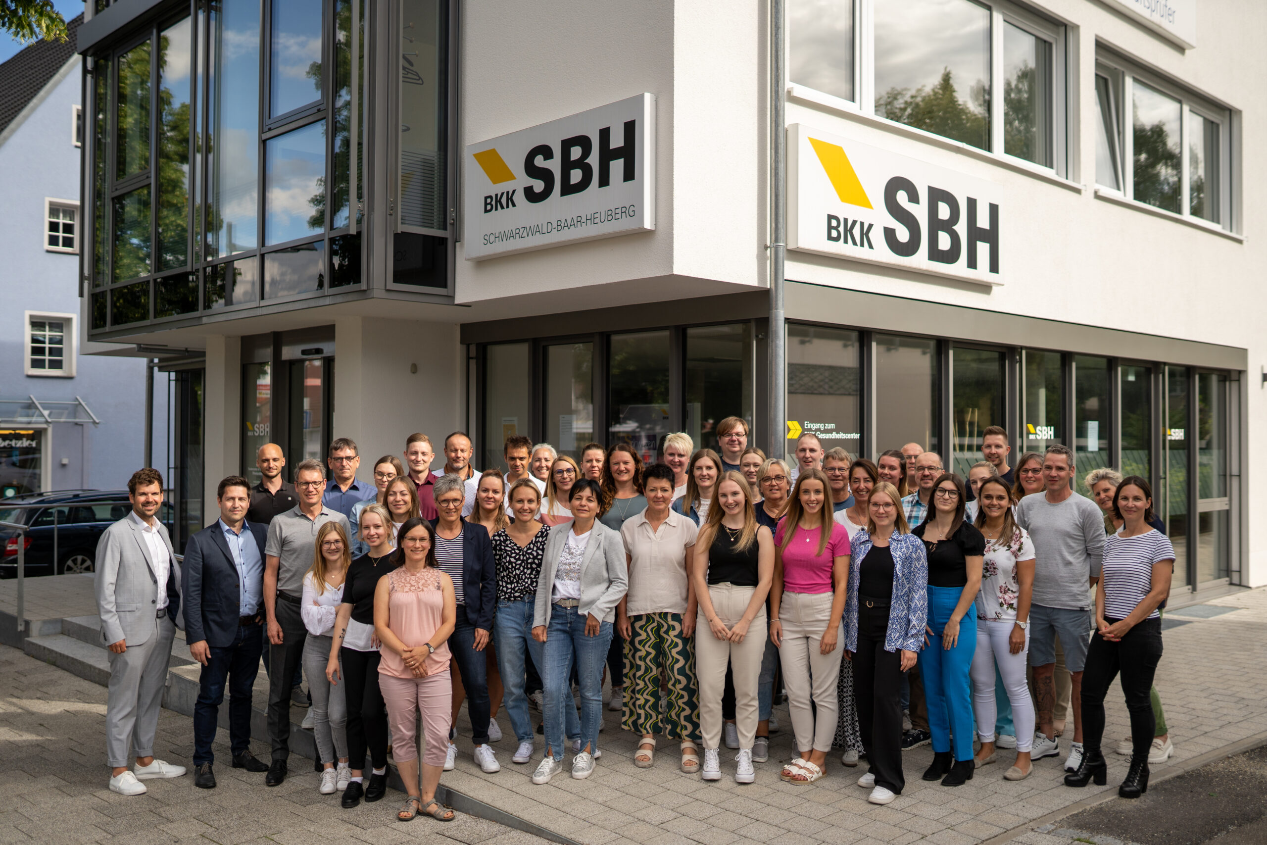 Team der BKK SBH vor einem Gebäude mit einem Schild BKK SBH Schwarzwald-Baar-Heuberg