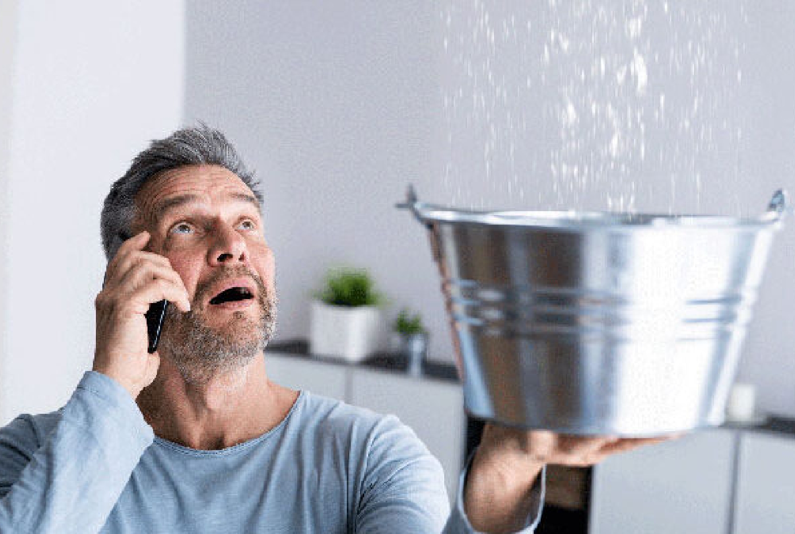 Mann mit einem erschrockenen Gesichtsausdruck und einer Hand mit Handy am rechten Ohr und der anderen Hand mit einem Eimer, der Wasser von oben auffängt