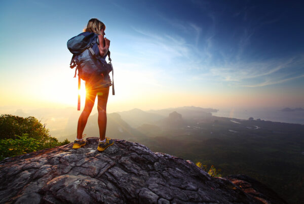 Frau mit einem Wanderrucksack steht auf einem Berg und beobachtet den Sonnenuntergang.