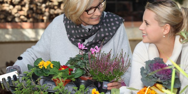 Eine ältere und eine jüngere Frau sitzen an einem Tisch. Vor ihnen stehen Pflanztöpfe mit Blumen, und eine Gartenharke liegt auf dem Tisch. Es scheint, als würden sie gemeinsam Blumen pflanzen.