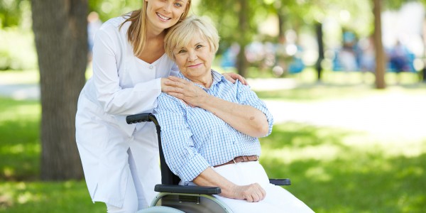 Eine herzliche Szene im Freien zeigt eine junge Frau, die sich liebevoll auf die Schultern einer älteren Frau im Rollstuhl lehnt. Die junge Frau, erkennbar als Pflegerin, strahlt Fürsorglichkeit aus, während die beiden gemeinsam die Natur genießen.