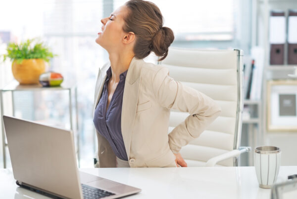 Eine Frau sitzt an ihrem Schreibtisch, hält sich ihren unteren Rücken fest und hat ein schmerzverzerrtes Gesicht.