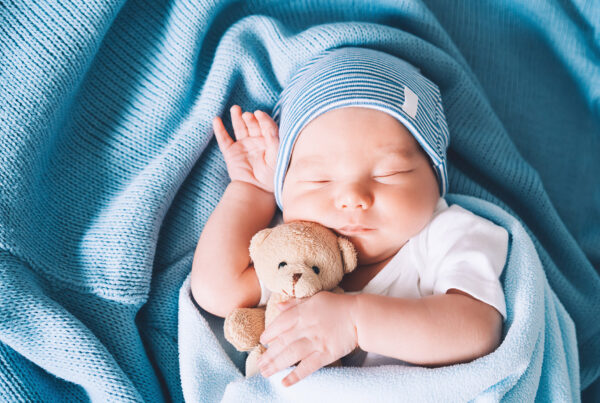 Ein schlafendes neugeborenes Baby, in eine blaue Decke eingewickelt, hält einen kleinen Teddybären in der Hand und trägt eine blau-weiß gestreifte Mütze auf dem Kopf.